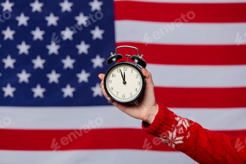 Preview: Female hand in Christmas sweater holds alarm clock on background with USA flag