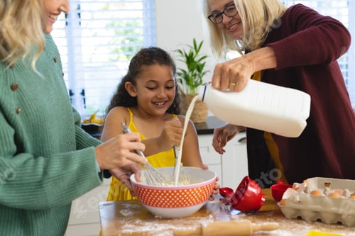 Preview: Multiracial senior woman pouring milk while mother and daughter whisking batter in bowl in kitchen