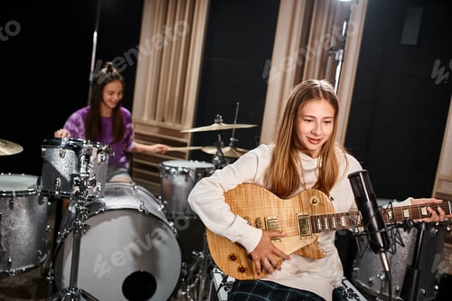 Visualização: concentre-se em uma linda guitarrista adolescente loira tocando ao lado de sua amiga desfocada tocando bateria no estúdio