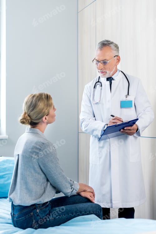 Preview: mature male doctor writing in clipboard near female patient in hospital room