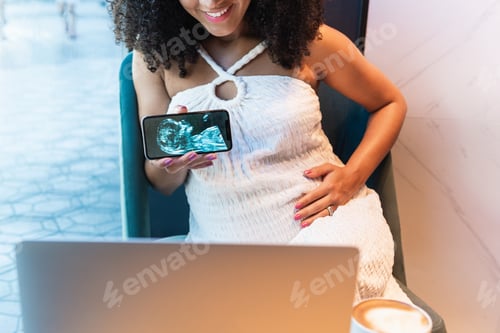 Preview: Anonymous black pregnant woman showing baby ultrasound picture to laptop