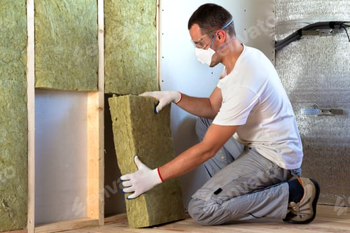 Preview: Worker in protective goggles and respirator insulating rock wool insulation in wooden frame for