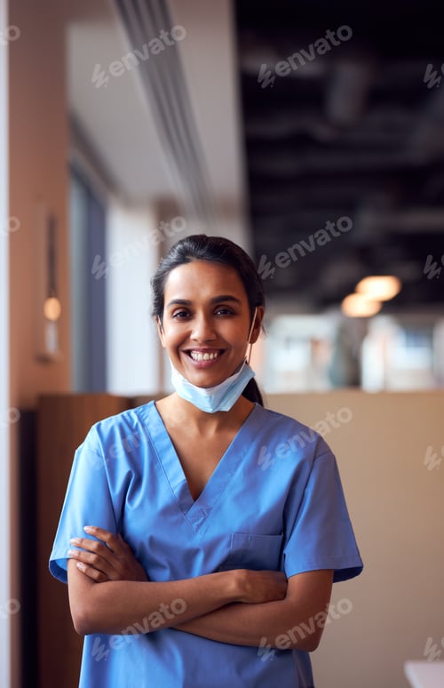 Preview: Smiling Female Doctor With Face Mask Wearing Scrubs In Busy Hospital During Health Pandemic