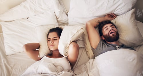 Preview: Shot of a young woman covering her ears with a pillow while her husband snores in bed