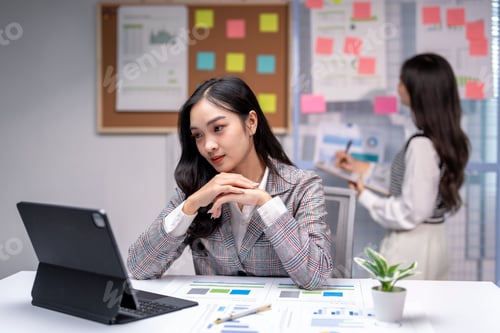 Preview: Businesswoman sitting at desk looking at tablet with coworker in background