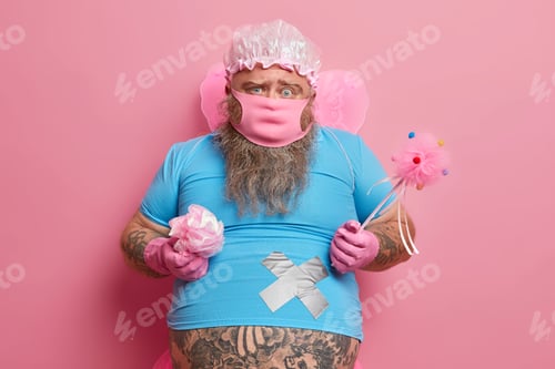 Preview: Cleaning concept. Displeased bearded plump man wears protective mask wings rubber gloves has tattooe