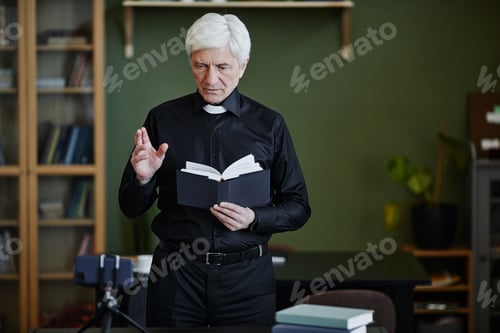 Preview: White haired senior priest holding Bible and recording video in office setting