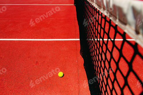 Preview: A tennis ball on the textured floor of a red court near the net after losing a match point.