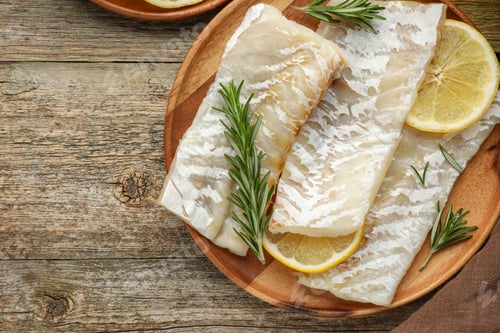 Preview: Fresh raw cod fillets with rosemary and lemon on wooden table, flat lay. Space for text