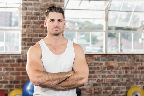 Preview: Muscular man standing arms crossed, wearing white tank top in gym with blue ball, yellow plates