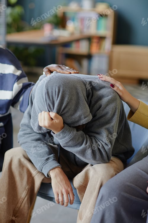 Preview: Teenage Boy Hiding Face in Hoodie Receiving Comfort from Support Group