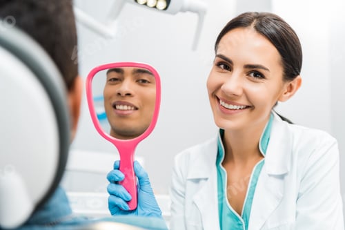 Preview: handsome african american man smiling while female dentist holding mirror