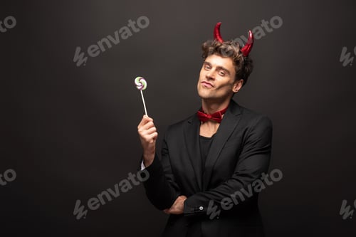 Preview: handsome man in halloween devil costume holding one lollipop on black