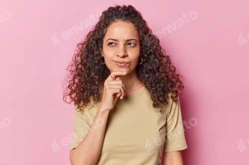 Vista previa: Portrait of thoughtful curly haired young woman keeps hand on chin focused aside contemplates about