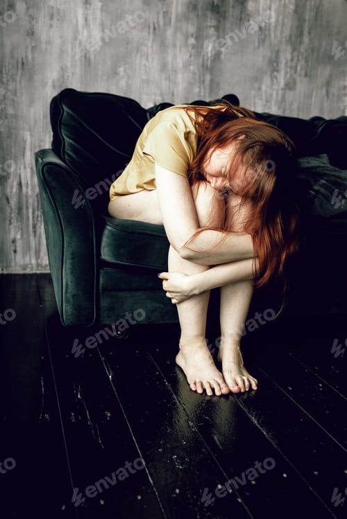 Preview: Young woman sitting on sofa, hugging her knees, her head down. Depression. Mental health.
