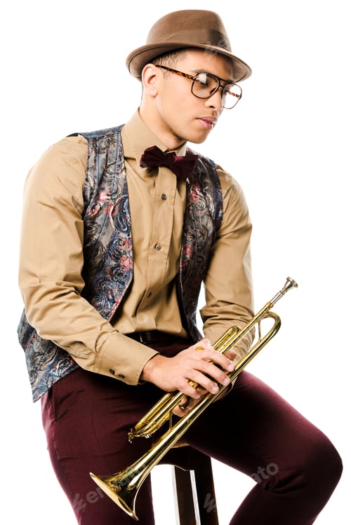 Preview: serious mixed race male jazzman holding trumpet and sitting on chair isolated on white
