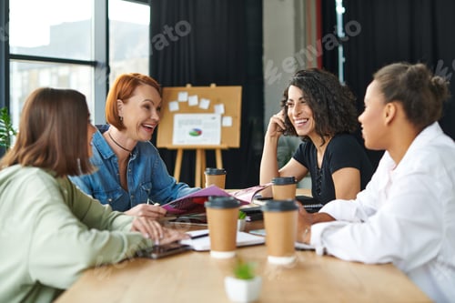 Preview: excited redhead woman holding magazine and laughing near takeaway drinks and diverse multicultural