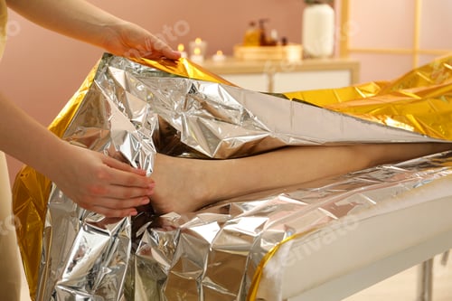 Preview: Spa body wraps. Esthetician covering woman with thermal foil blanket indoors, closeup