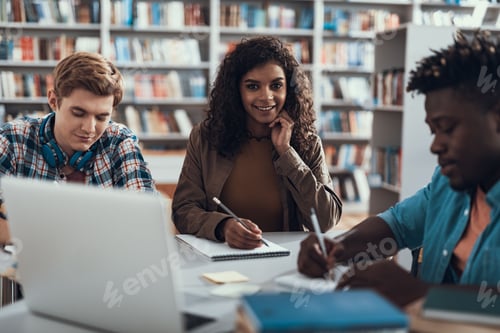 Preview: Attractive Long Haired Student Sitting And Smiling While Two Young Men Doing Their Homework