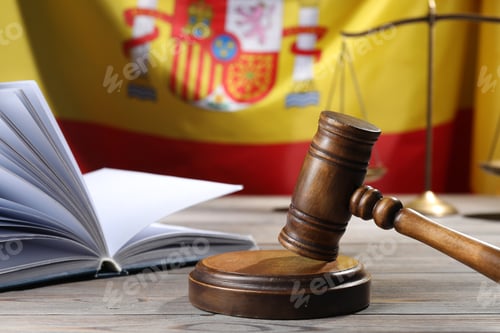 Preview: Judge's gavel, book and scales on wooden table against flag of Spain, closeup