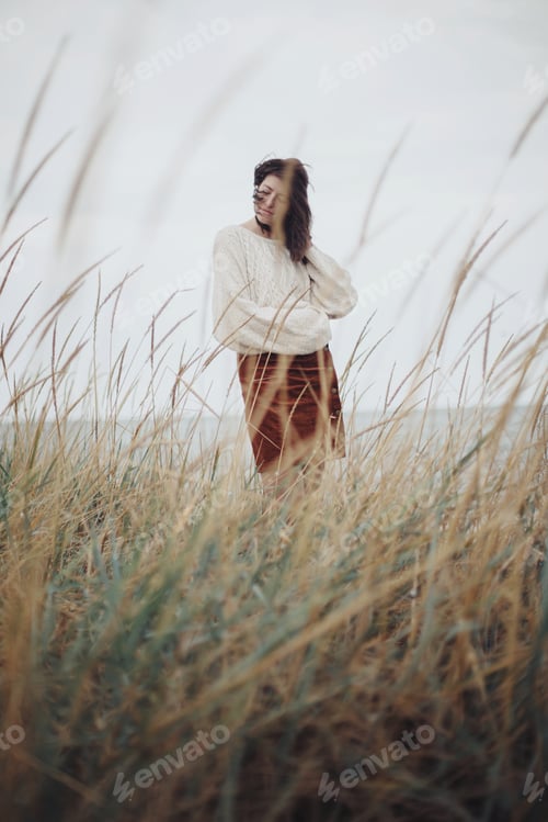 Preview: Beautiful stylish woman with windy hair in knitted sweater posing among wild grass. Tranquility