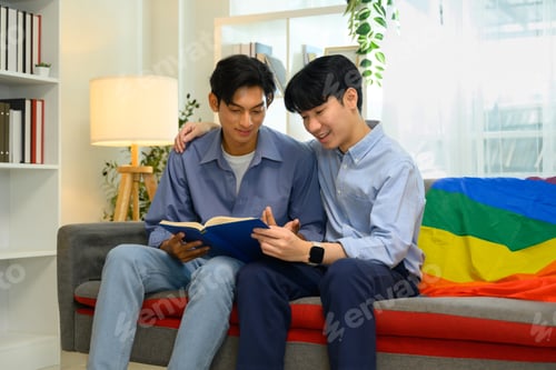 Preview: Happy male gay couple enjoying quality time together and reading book on couch