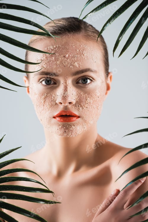 Preview: naked woman with makeup and scrub on face looking at camera near palm leaves on white