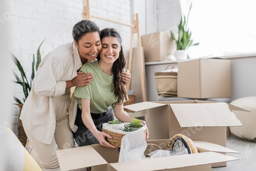Preview: cheerful multiracial woman hugging shoulders of happy girlfriend while moving to new house