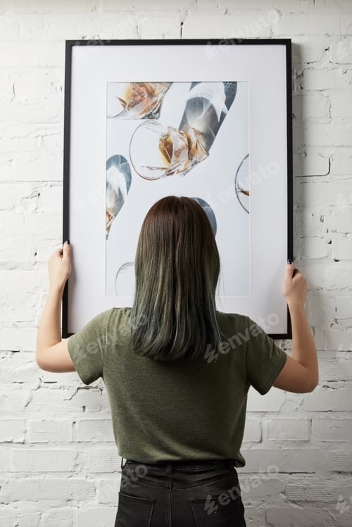 Preview: back view of woman holding drawing in black square frame in hands