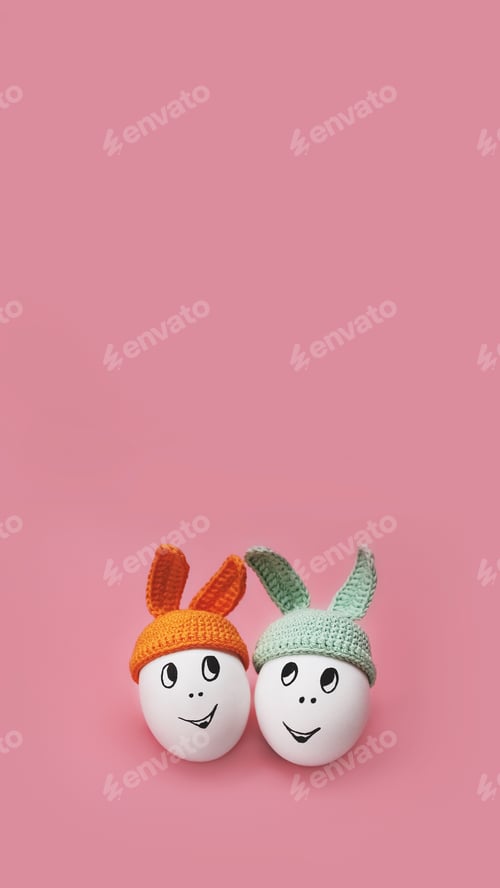 Preview: two white eggs in beanie with rabbit ears and cheerful faces on pink background, vertical, 16:9