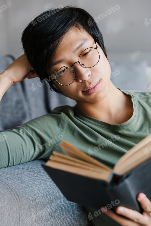 Preview: Image of young asian man wearing eyeglasses reading book in apartment