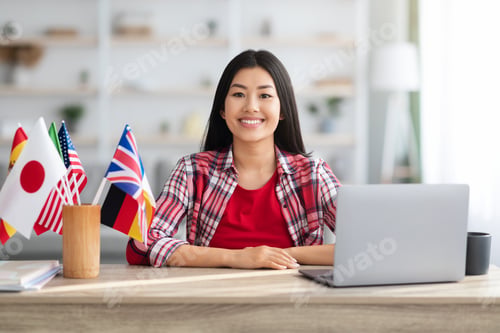 Preview: Smiling Asian Lady Sitting At Table With International Flags And Laptop