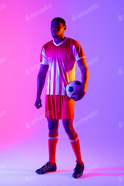 Preview: Portrait of african american male soccer player with football over neon pink lighting