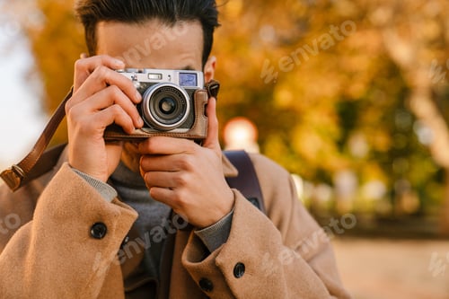 Preview: Young man wearing coat taking photo on retro camera in autumn park
