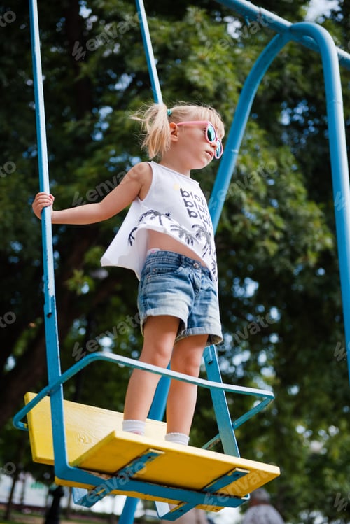 Preview: Stylish little girl in pink sunglasses swinging on playground