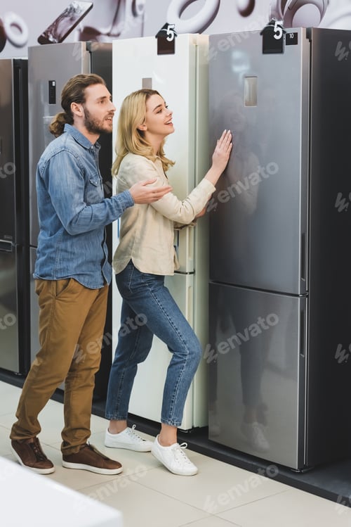 Preview: boyfriend pointing with hand and smiling girlfriend touching fridge in home appliance store