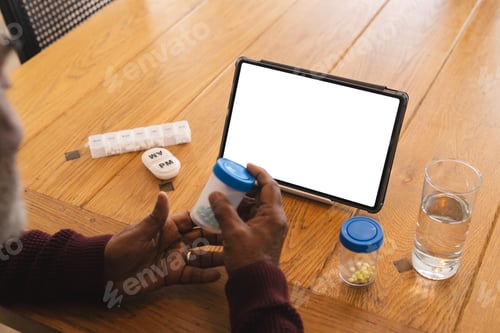 Preview: Senior biracial man having video call with doctor on tablet with copy space, holding pills at home