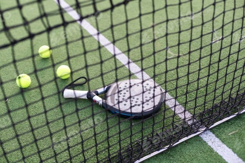 Preview: Blue padel racket and yellow balls placed on court near net on sunny day