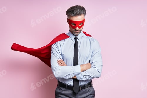Preview: Confident mature man in shirt and tie wearing superhero cape while standing against pink background