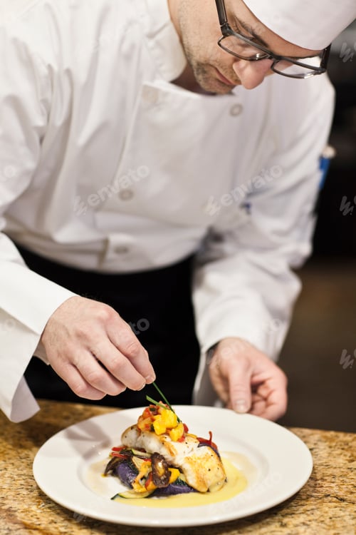 Preview: A caucasian male chef putting the finishing touches on a plate of fish in a commercial kitchen.