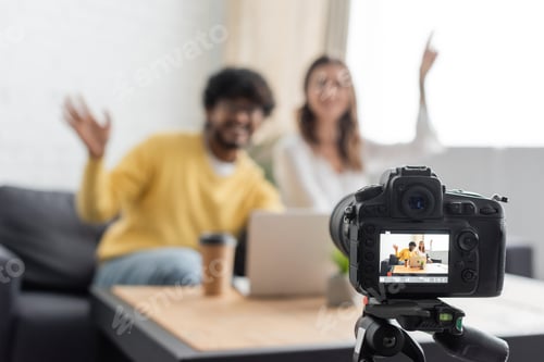 Preview: selective focus of professional digital camera near blurred interracial vloggers waving hands near