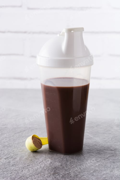 Preview: Whey protein chocolate shake in shaker bottle with scoop on grey table