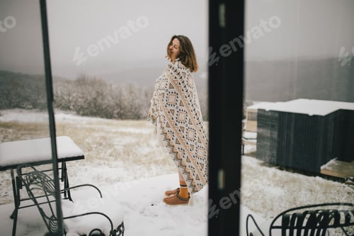 Vista previa: Mujer joven envuelta en una cálida manta de pie en un balcón con un bosque nevado al fondo.