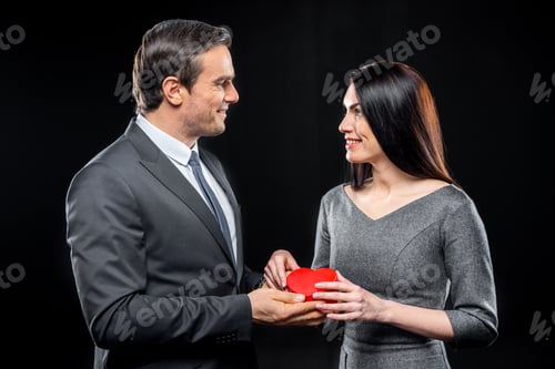 Preview: Couple holding red toy heart smiling and looking at each other isolated on black