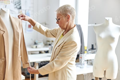 Preview: Middle aged woman designer measures sleeve of elegant jacket in light workshop