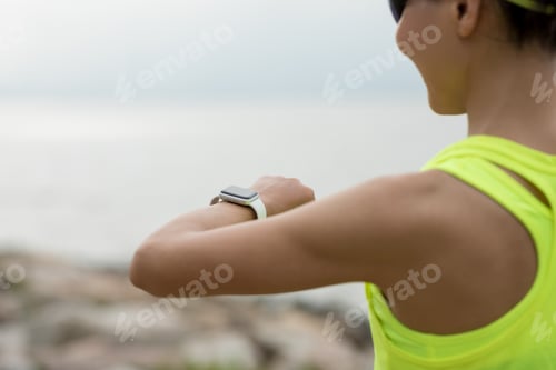 Preview: Fitness woman runner looking at her smartwatch on sunny coast trail