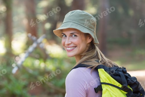 Preview: Woman standing on wooded trail turning to smile with bucket hat, yellow-black backpack, copy space