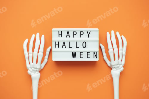 Preview: HAPPY HALLOWEEN inscription on lightbox on orange background with skeleton hands.