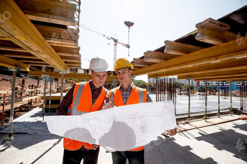 Preview: Two men dressed in shirts, orange work vests and helmets explore construction documentation on the