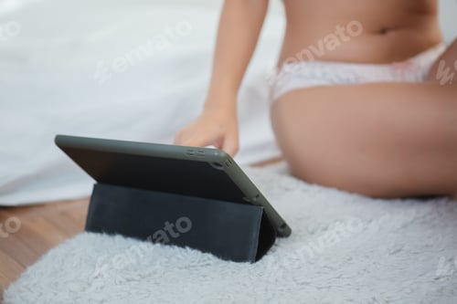 Preview: Young woman in underwear sitting using digital tablet at home in bedroom.